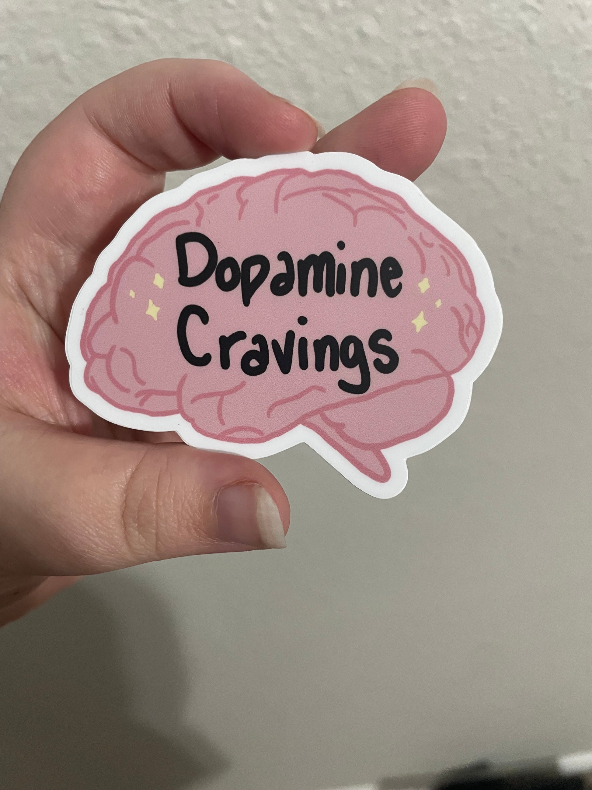 Brain shaped pink sticker that says Dopamine Cravings in black writing. There are small yellow sparkle icons next to either side of the words.